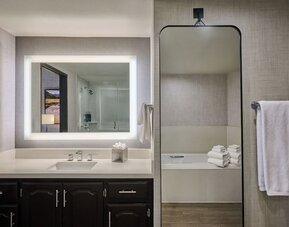 Modern hotel bathroom with a wide illuminated vanity mirror, white countertop and sink, dark wood cabinetry, towel rack, and a bathtub visible through a tall wall mirror.