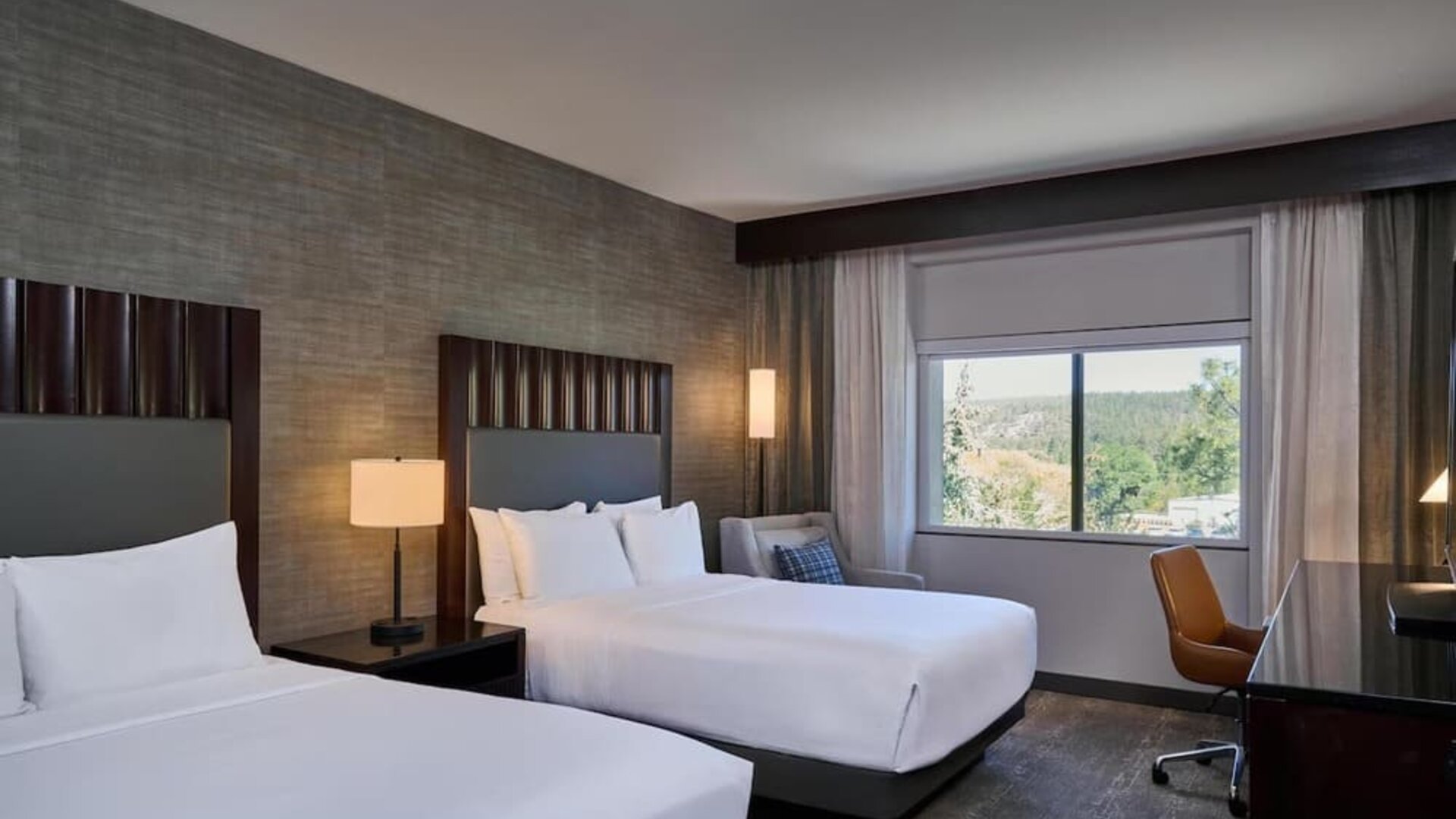 Modern hotel guest room with two beds dressed in white linens, dark upholstered headboards, bedside lighting, a work desk and chair, and a large window overlooking a wooded landscape.