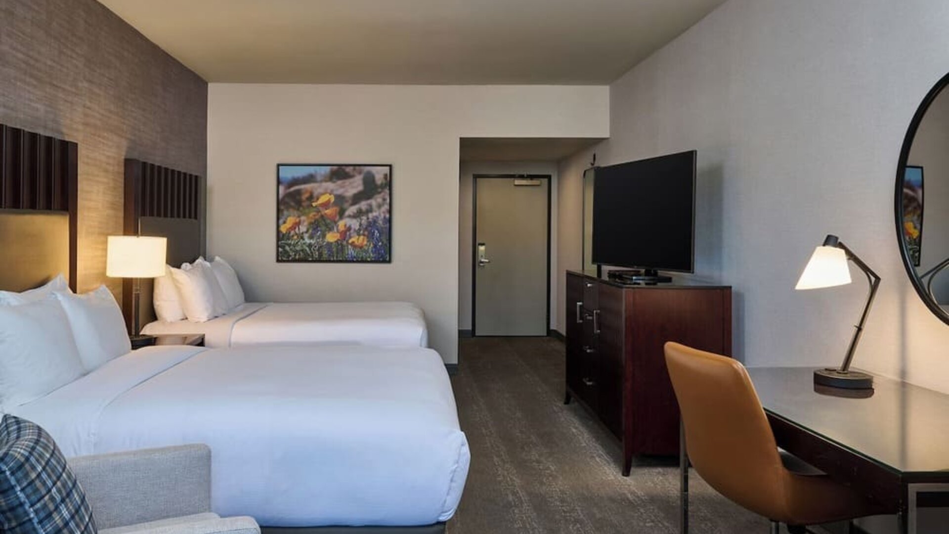 Spacious hotel guest room with two beds in white linens, dark headboards, an upholstered armchair, flat-screen television on a dresser, and a desk with lamp, chair, and round wall mirror.