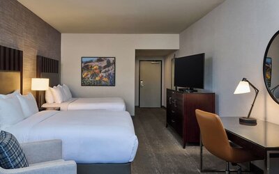 Spacious hotel guest room with two beds in white linens, dark headboards, an upholstered armchair, flat-screen television on a dresser, and a desk with lamp, chair, and round wall mirror.