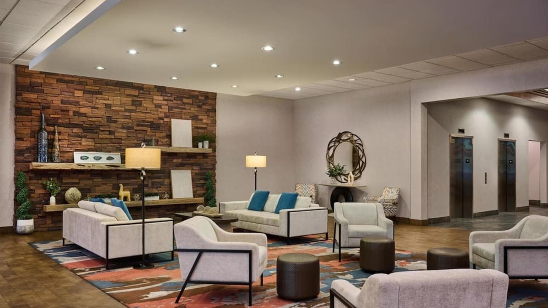 Contemporary hotel lobby seating area with cream sofas and armchairs, teal accent pillows, colorful patterned rug, floor lamps, stone accent wall, decorative shelving, and elevators nearby.