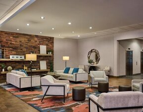 Contemporary hotel lobby seating area with cream sofas and armchairs, teal accent pillows, colorful patterned rug, floor lamps, stone accent wall, decorative shelving, and elevators nearby.