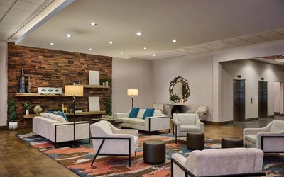 Contemporary hotel lobby seating area with cream sofas and armchairs, teal accent pillows, colorful patterned rug, floor lamps, stone accent wall, decorative shelving, and elevators nearby.