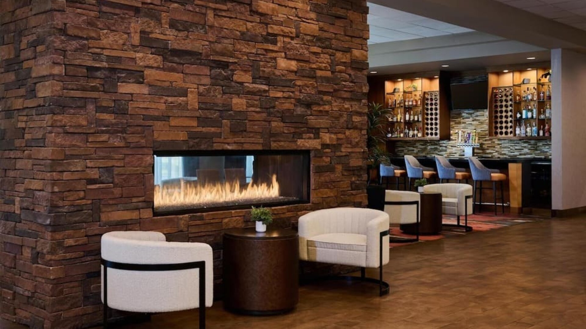 Cozy hotel lounge featuring a large stacked-stone fireplace with visible flames, two cream armchairs and a side table, with a warmly lit bar and counter seating in the background.