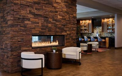 Cozy hotel lounge featuring a large stacked-stone fireplace with visible flames, two cream armchairs and a side table, with a warmly lit bar and counter seating in the background.