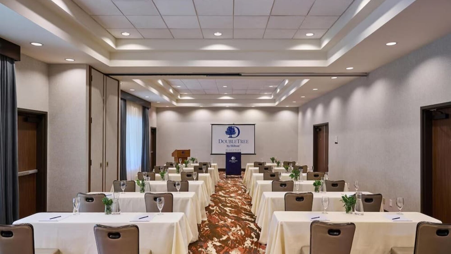 Spacious hotel meeting room arranged classroom-style with rows of white-covered tables and chairs, water glasses, small plants, a podium, and a DoubleTree by Hilton presentation screen.
