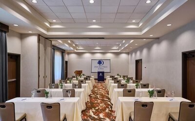 Spacious hotel meeting room arranged classroom-style with rows of white-covered tables and chairs, water glasses, small plants, a podium, and a DoubleTree by Hilton presentation screen.