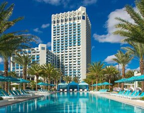 Palm-lined resort swimming pool with turquoise water, rows of blue lounge chairs and umbrellas, private cabanas, and a tall white hotel tower rising in the background.