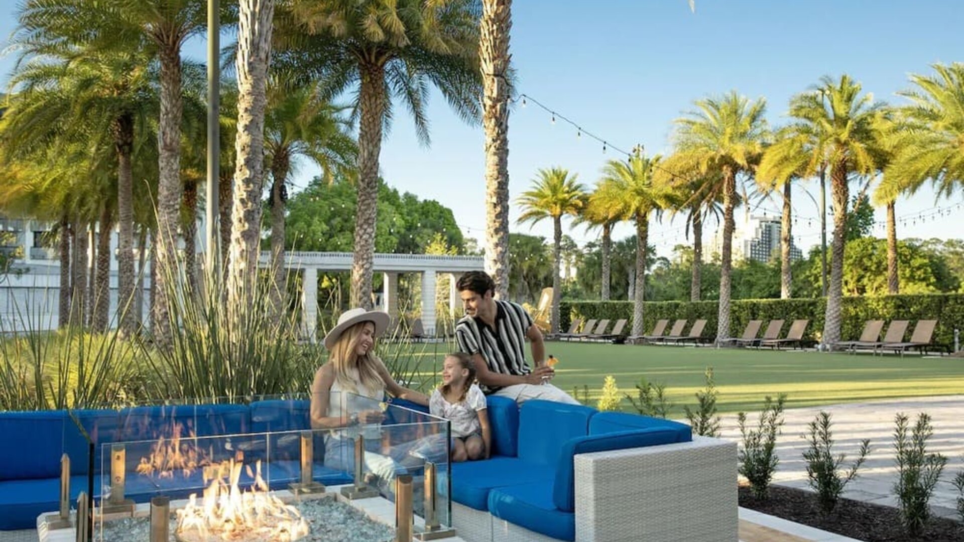 Outdoor resort seating area with bright blue cushioned sofas surrounding a glass-enclosed fire pit, framed by palm trees, landscaped grounds, string lights, and rows of lounge chairs.