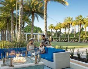 Outdoor resort seating area with bright blue cushioned sofas surrounding a glass-enclosed fire pit, framed by palm trees, landscaped grounds, string lights, and rows of lounge chairs.