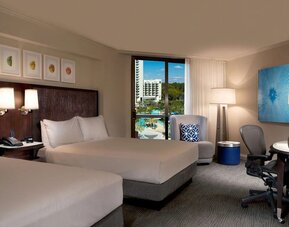 Spacious hotel room with two beds dressed in white linens, dark headboard with reading lights, armchair, work desk and ergonomic chair, blue accent decor, and a window overlooking the resort pool area.