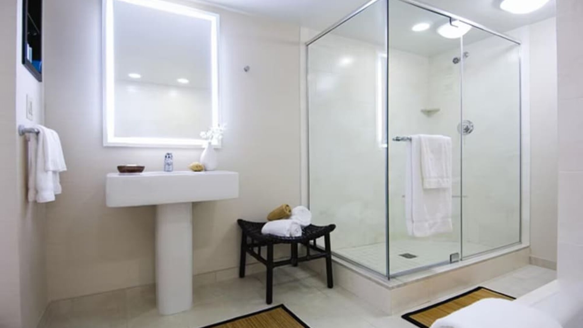 Bright hotel bathroom with illuminated vanity mirror, pedestal sink, glass-enclosed walk-in shower, white towels, bench, neutral tile flooring, and simple contemporary decor.