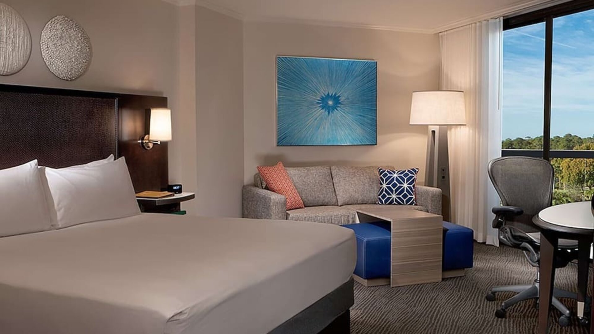 Contemporary king guest room with white bedding, dark headboard, sofa, blue ottomans, work desk and ergonomic chair, floor lamp, colorful wall art, and a large window with a treetop view.