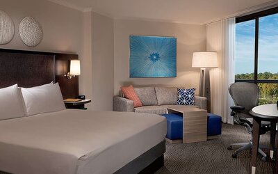 Contemporary king guest room with white bedding, dark headboard, sofa, blue ottomans, work desk and ergonomic chair, floor lamp, colorful wall art, and a large window with a treetop view.