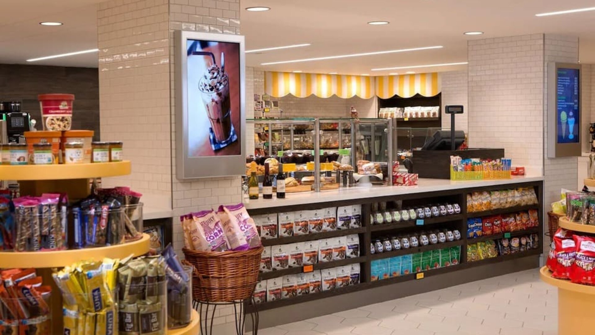 Bright hotel market with shelves and displays stocked with snacks, drinks, packaged foods, and grab-and-go items, plus a service counter and refrigerated cases.