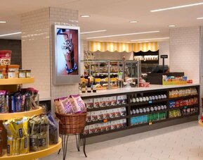 Bright hotel market with shelves and displays stocked with snacks, drinks, packaged foods, and grab-and-go items, plus a service counter and refrigerated cases.