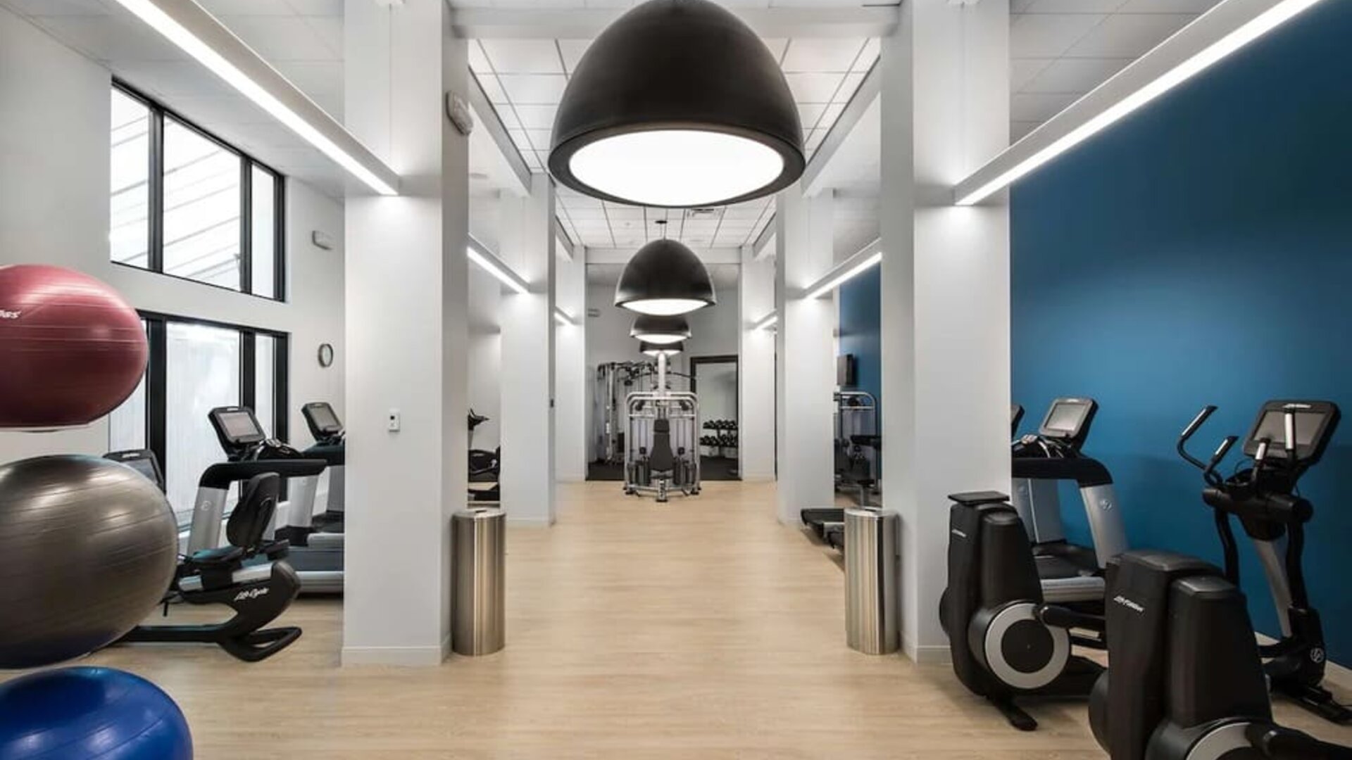 Spacious hotel fitness center with treadmills, elliptical machines, strength-training equipment, exercise balls, and weights beneath oversized pendant lights and a bright contemporary interior.