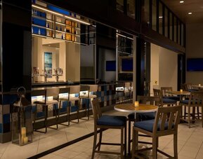 Contemporary hotel bar with illuminated bottle displays, counter seating, high-top tables and chairs, televisions, decorative lanterns, and warm ambient lighting.