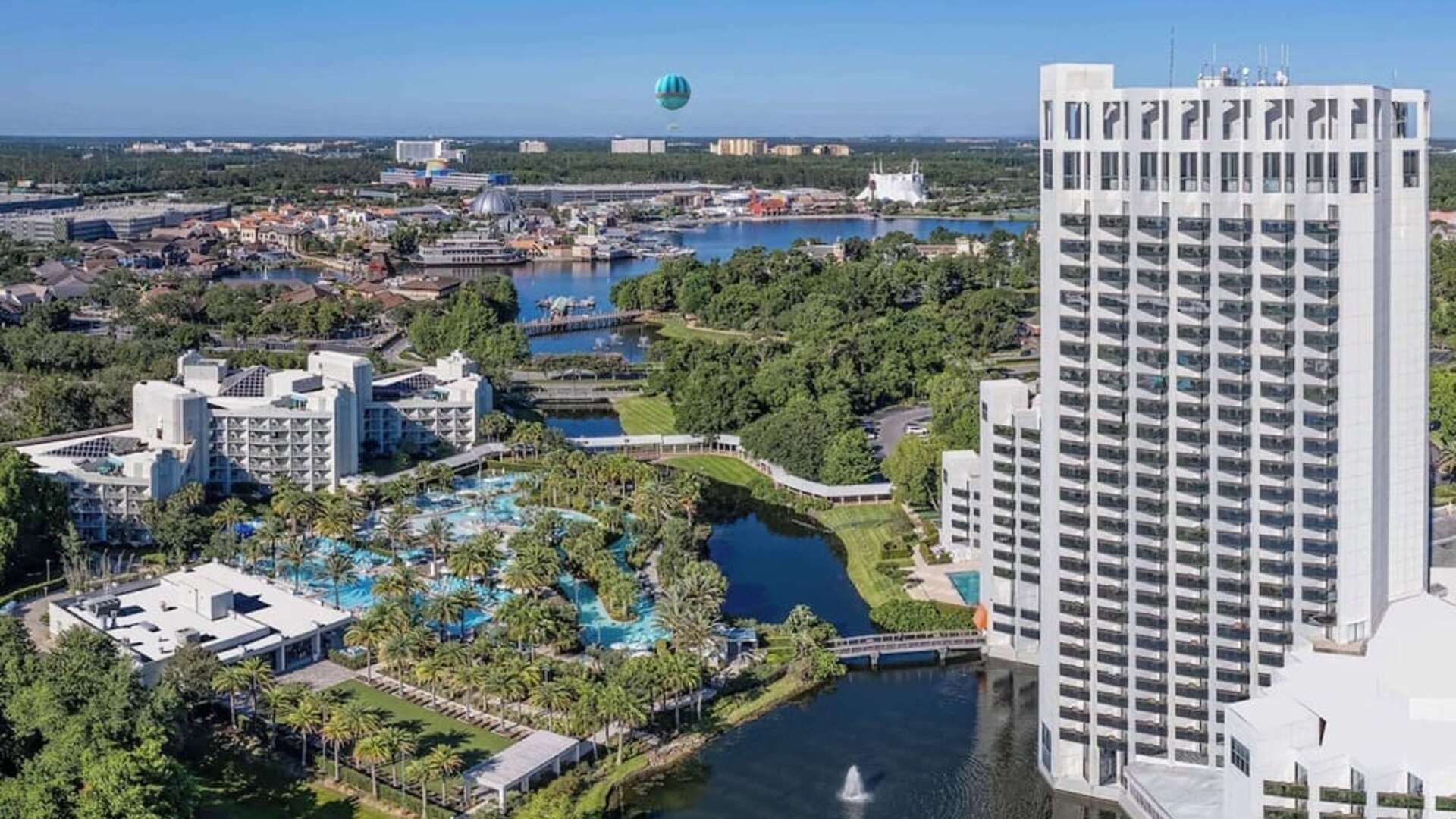 Aerial view of a tall white resort tower beside landscaped grounds, winding waterways, palm-lined swimming pools, and nearby buildings extending across the surrounding area.