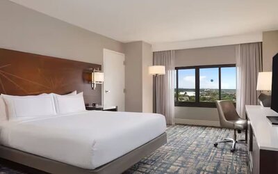 Spacious hotel room with king bed, white linens, large window with an elevated outdoor view, work desk and chair, flat-screen TV, bedside lighting, and patterned carpet.