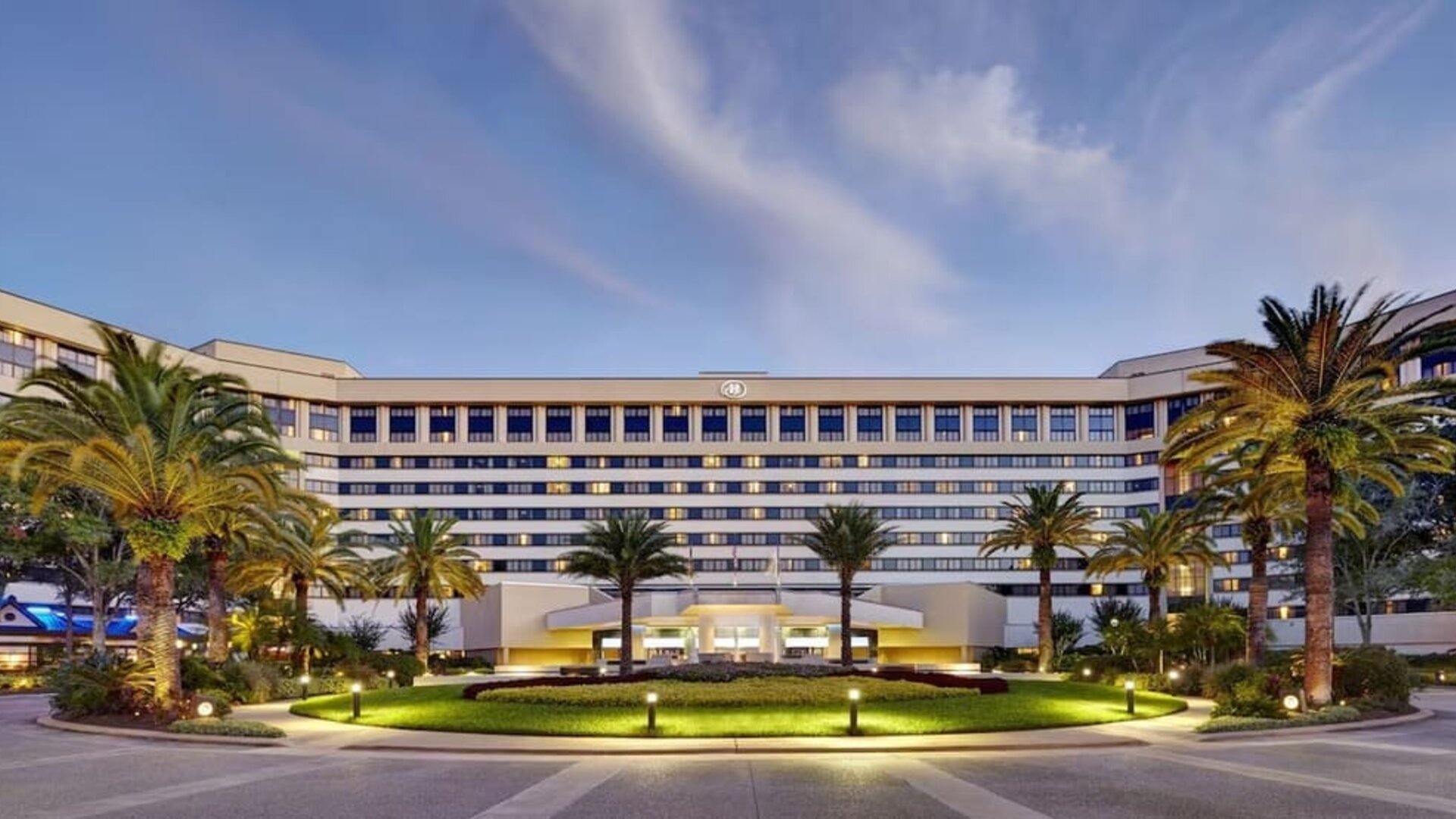 Large multi-story hotel exterior at dusk with illuminated windows and entrance, landscaped circular lawn, mature palm trees, pathways, and exterior lighting.