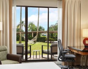 Hotel guest room with desk and ergonomic chair beside sliding glass doors opening to a furnished patio, green lawn, palm trees, and a hammock.