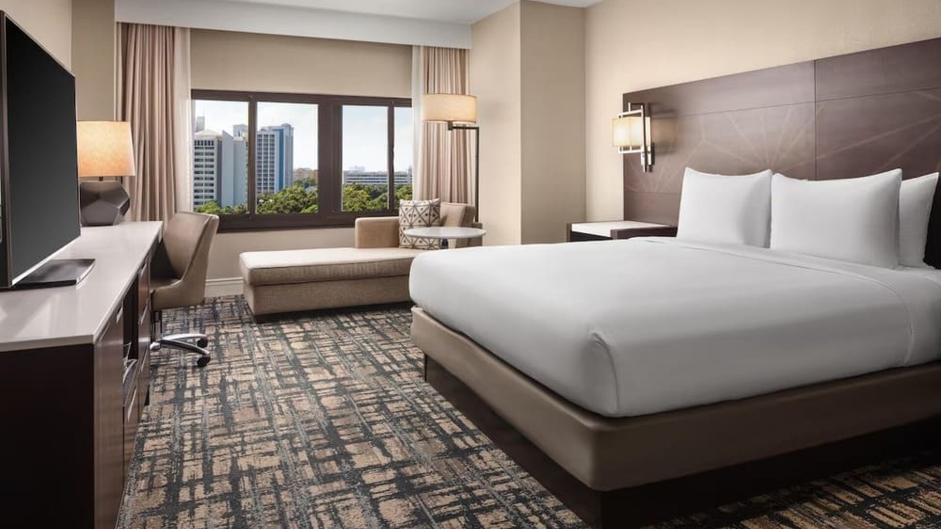 Contemporary hotel room with king bed in crisp white linens, large flat-screen TV, work desk and chair, chaise lounge, patterned carpet, and window overlooking nearby buildings and greenery.