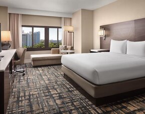 Contemporary hotel room with king bed in crisp white linens, large flat-screen TV, work desk and chair, chaise lounge, patterned carpet, and window overlooking nearby buildings and greenery.