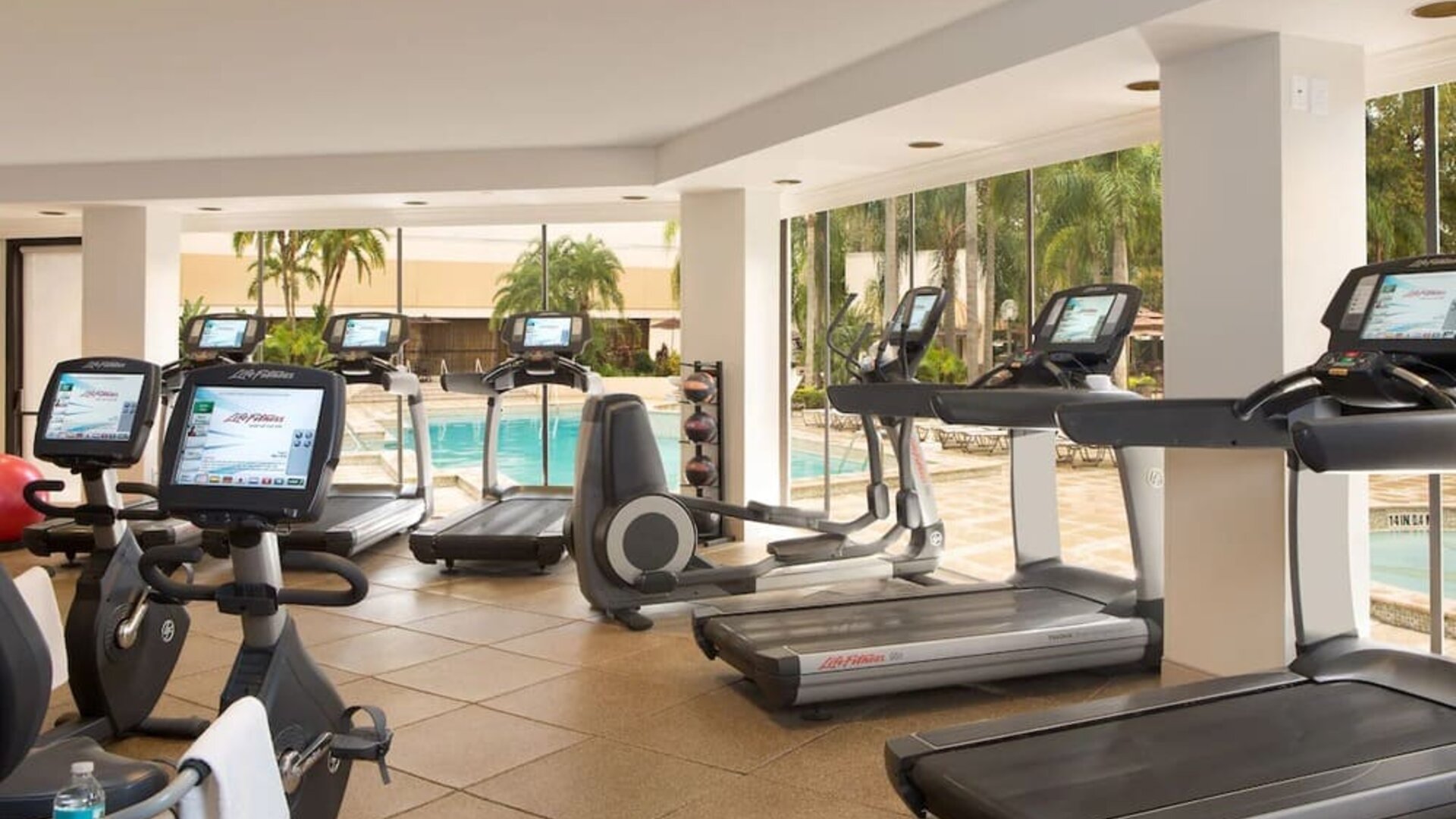 Bright hotel fitness center with treadmills, stationary bikes, elliptical machines, and other cardio equipment overlooking an outdoor swimming pool through large windows.