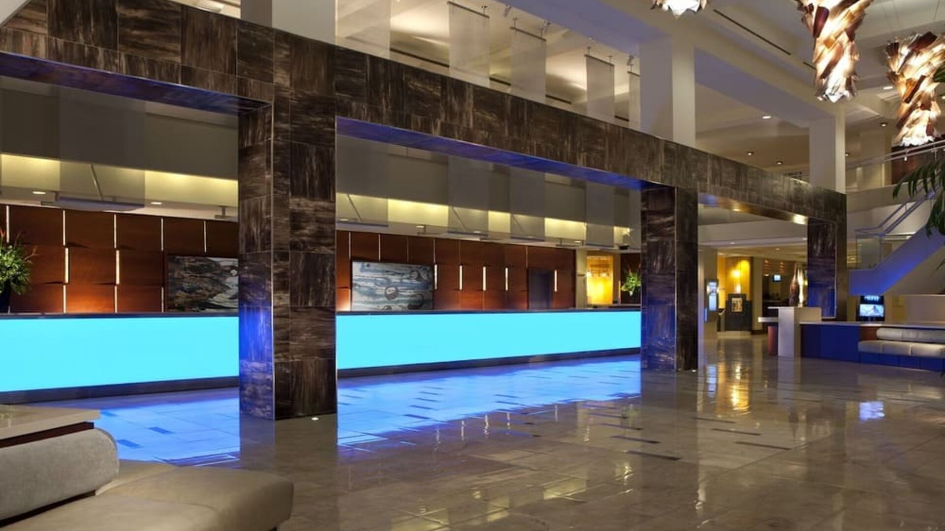 Contemporary hotel lobby with long illuminated blue reception counters, glossy tile flooring, dark architectural columns, modern hanging lights, and seating areas.