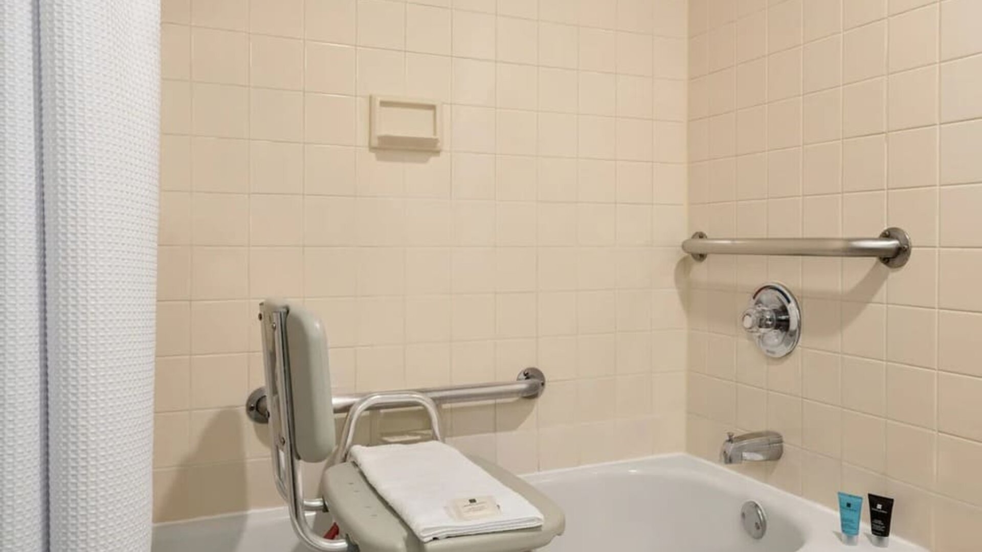 Accessible bathroom featuring a bathtub with transfer bench, grab bars, shower curtain, and bath amenities. Crowne Plaza Englewood.