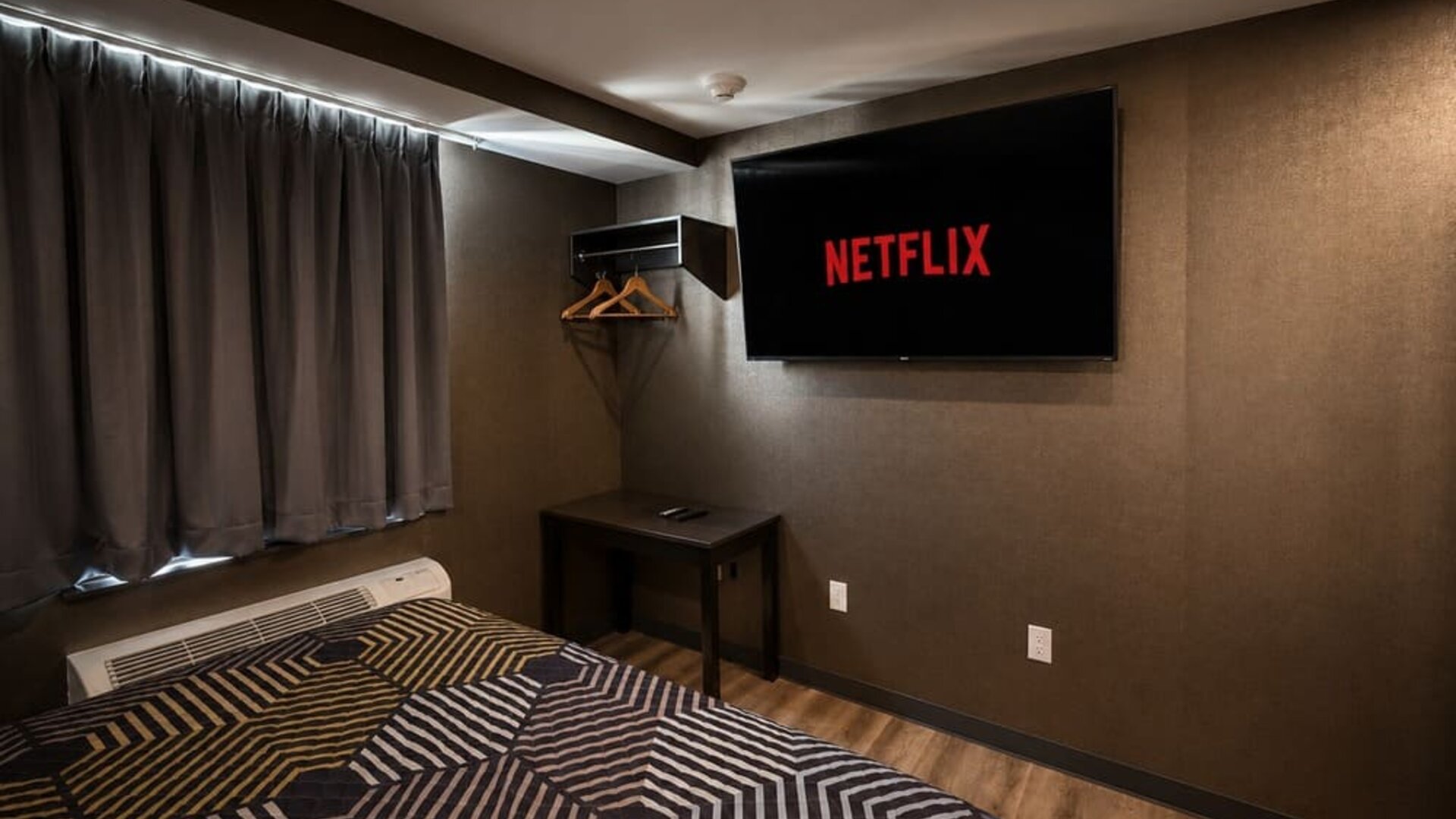 Dark contemporary guest room with geometric-patterned king bed, wall-mounted television displaying Netflix, blackout curtains, open hanging rack, and small table.
