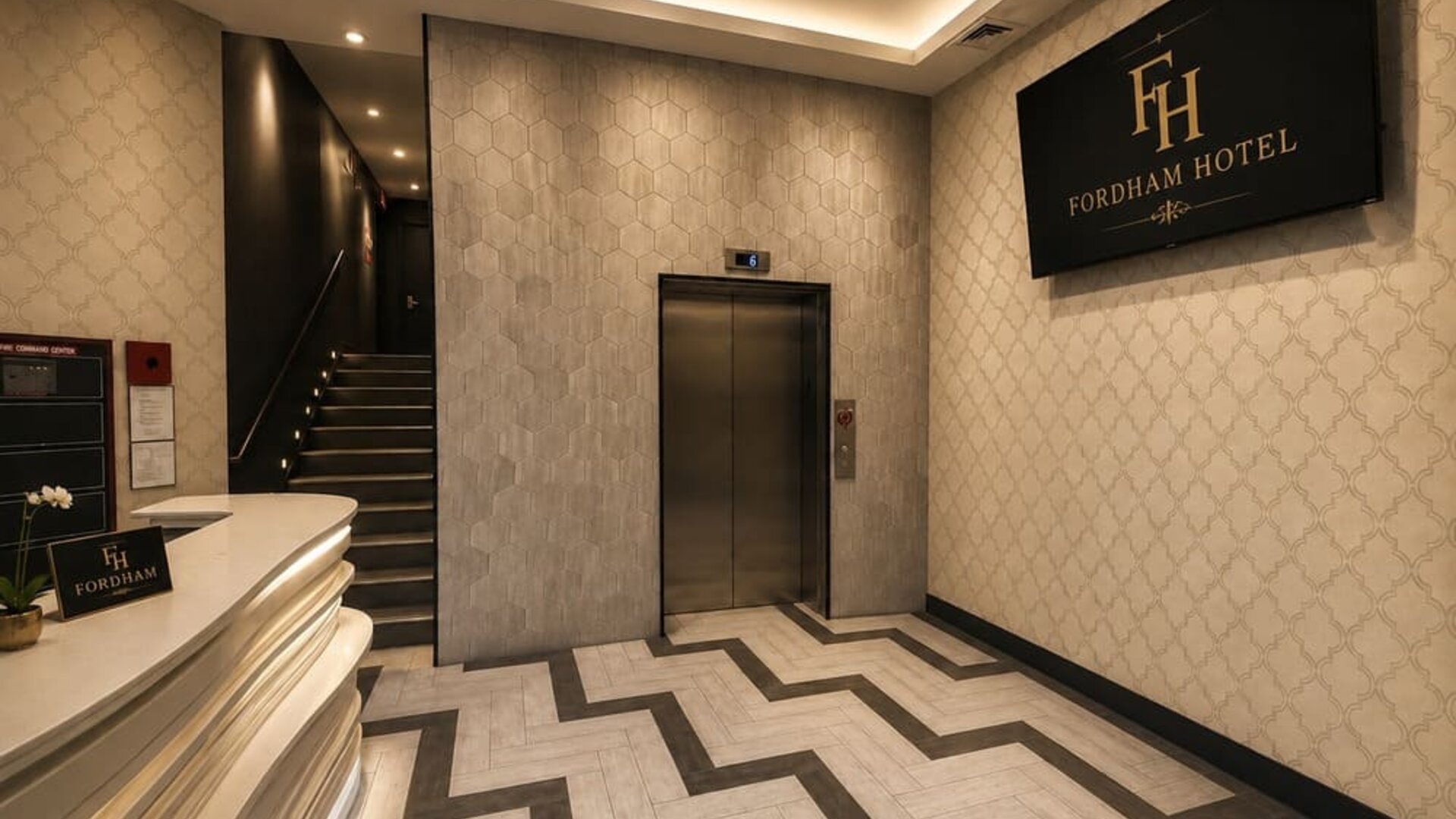 Fordham Hotel lobby with curved white reception desk, elevator, illuminated staircase, patterned tile flooring, decorative walls, and large hotel sign.