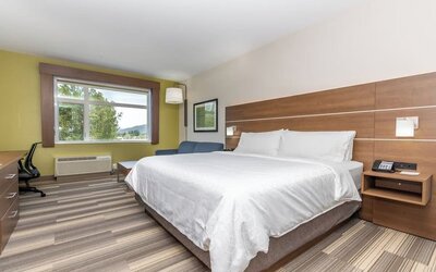 Spacious hotel guest room with a king-size bed, white linens, wood headboard, bedside table, work area, seating, and large window with an outdoor view.