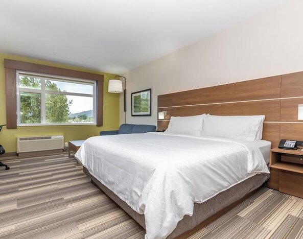 Spacious hotel guest room with a king-size bed, white linens, wood headboard, bedside table, work area, seating, and large window with an outdoor view.
