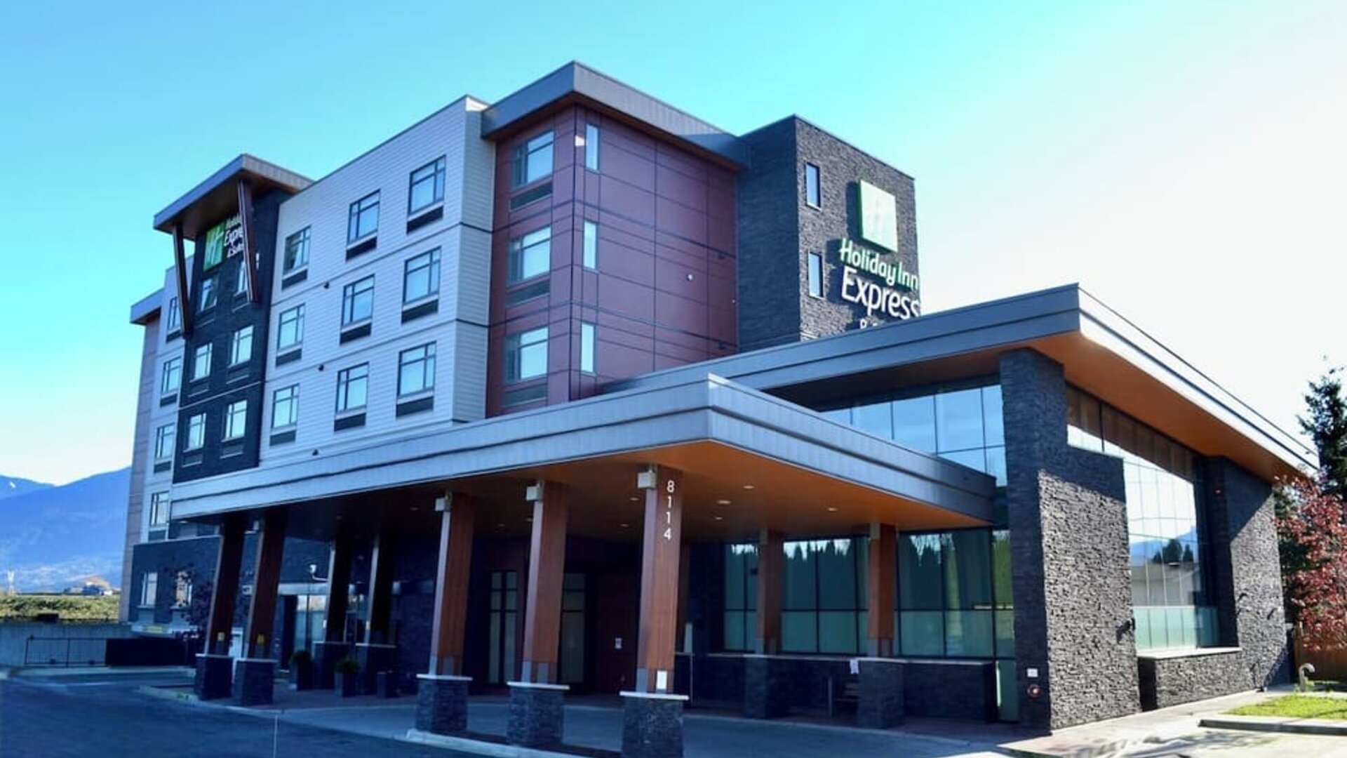 Modern Holiday Inn Express hotel exterior with a multi-story façade, covered entrance, large windows, stone accents, and mountain scenery in the distance.