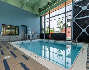 Indoor hotel swimming pool with a tiled deck, floor-to-ceiling windows, accessibility lift, safety equipment, and contemporary high-ceiling design.