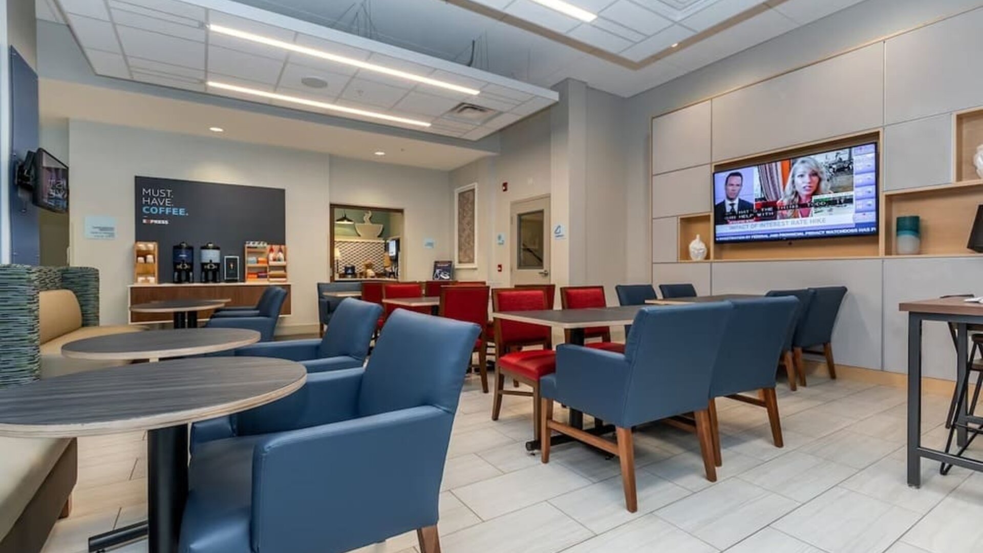 Contemporary hotel dining and lounge area with blue and red seating, round tables, beverage station, wall-mounted televisions, and modern lighting.