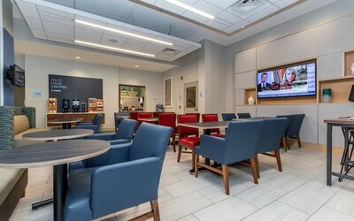 Contemporary hotel dining and lounge area with blue and red seating, round tables, beverage station, wall-mounted televisions, and modern lighting.