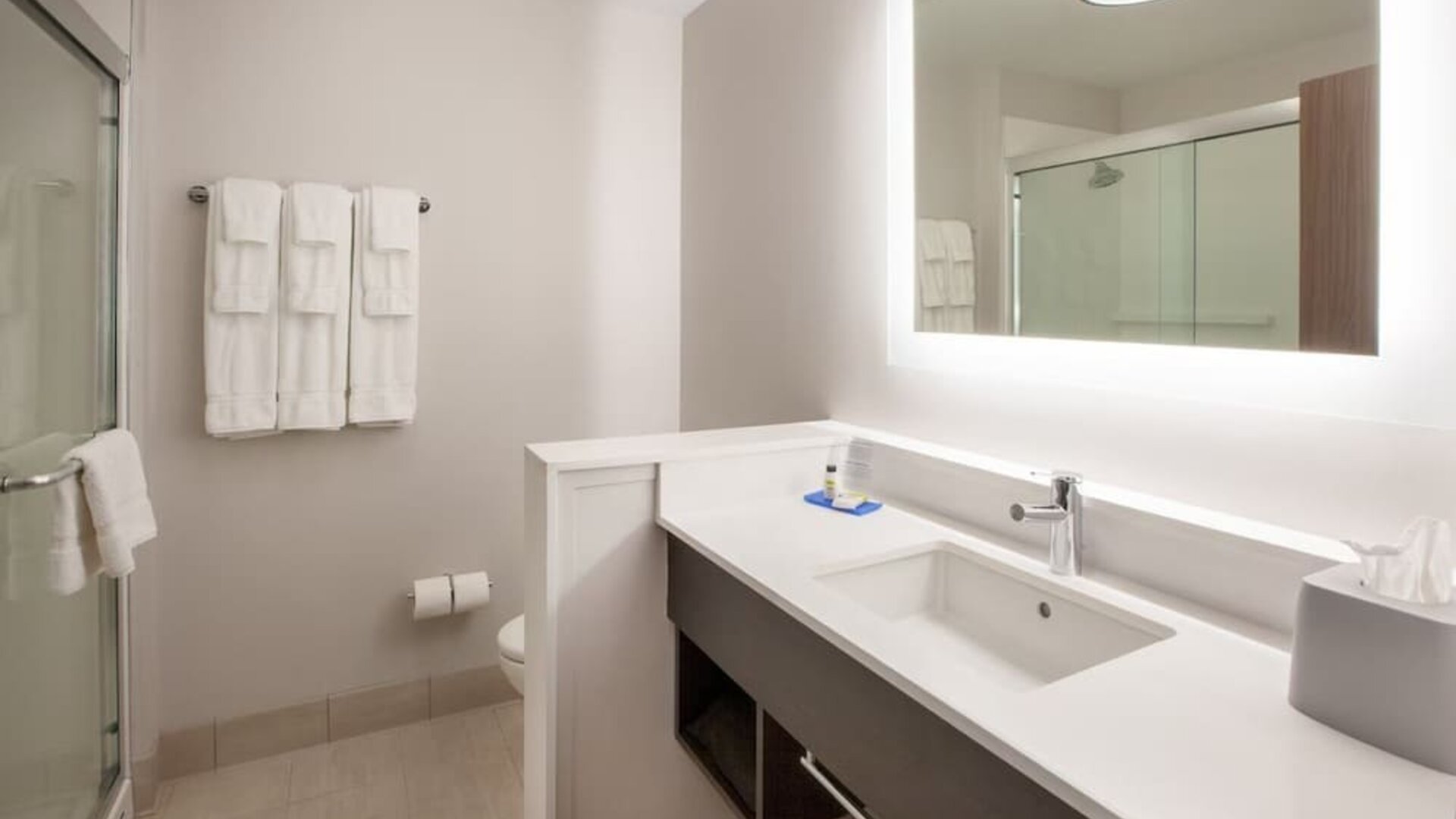 Bright hotel bathroom with a large illuminated mirror, white vanity and sink, glass-enclosed shower, toilet, and neatly arranged white towels.