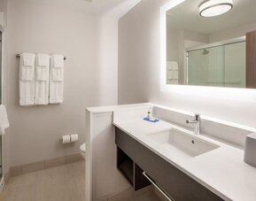 Bright hotel bathroom with a large illuminated mirror, white vanity and sink, glass-enclosed shower, toilet, and neatly arranged white towels.