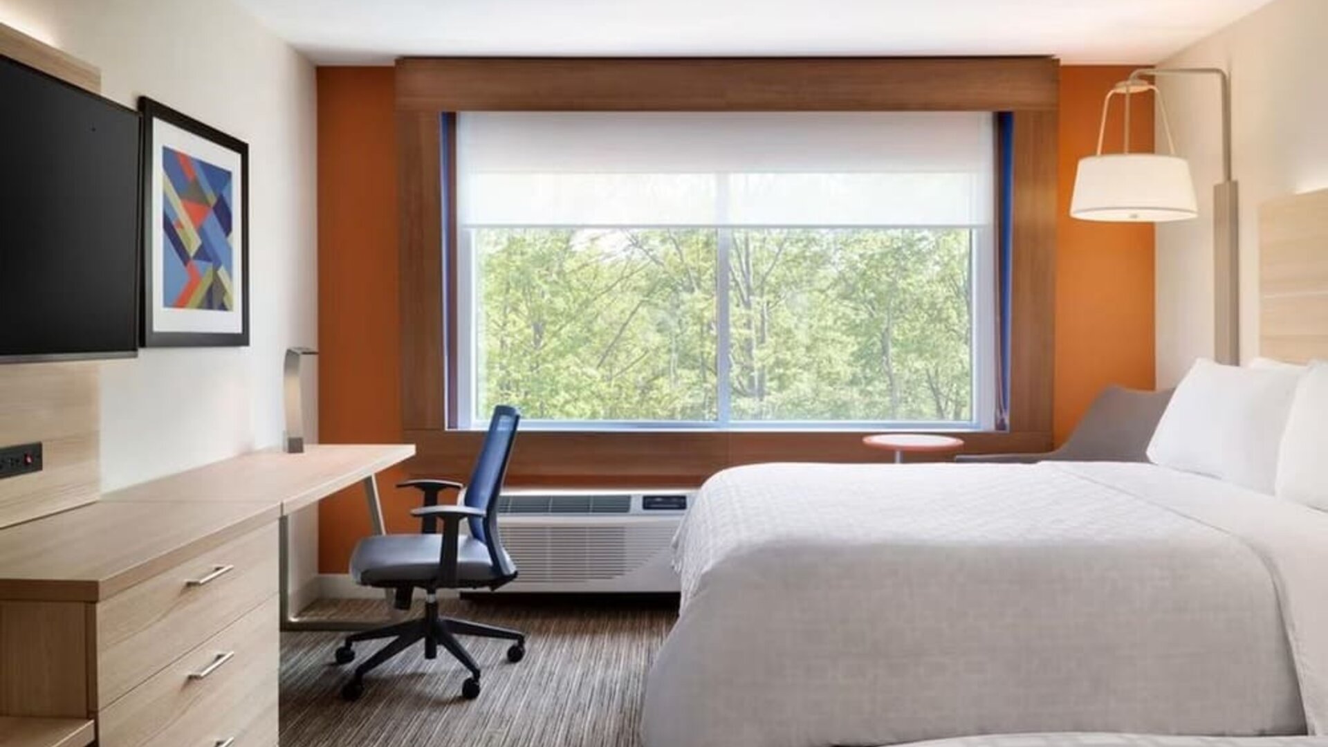 Bright, modern hotel room with two white beds, a work desk and chair, wall-mounted TV, and large window overlooking green trees.