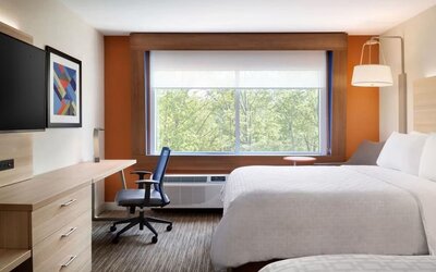 Bright, modern hotel room with two white beds, a work desk and chair, wall-mounted TV, and large window overlooking green trees.