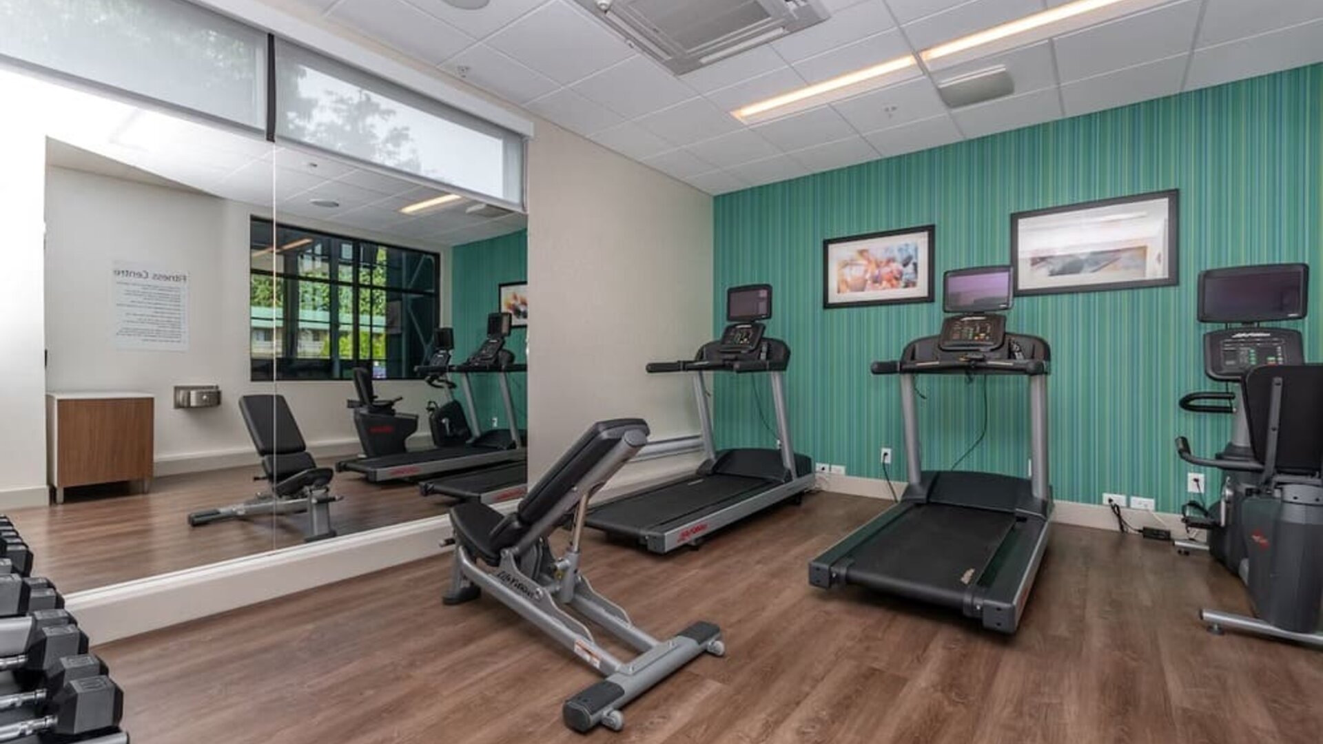 Modern hotel fitness center with treadmills, exercise bikes, adjustable weight bench, dumbbells, mirrored wall, and wood-look flooring.