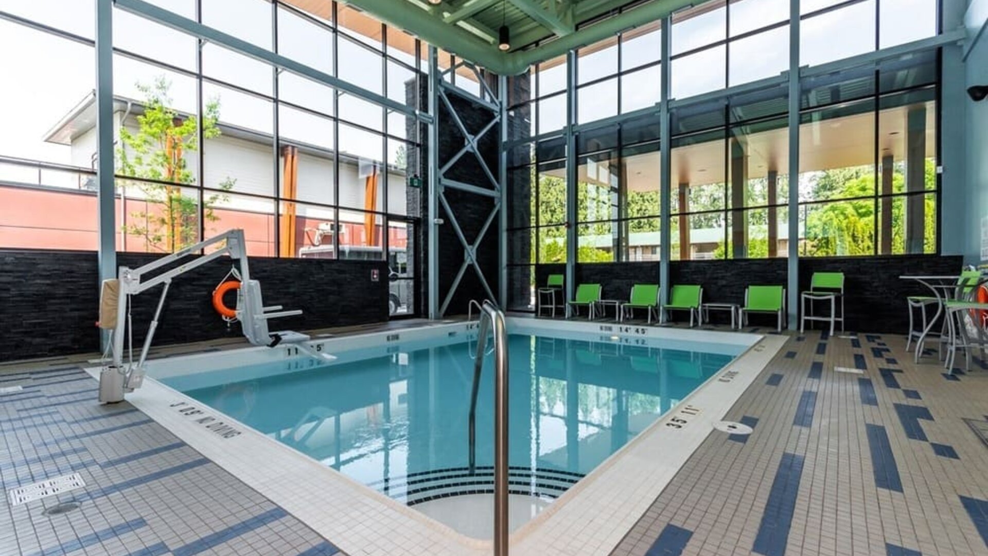 Bright indoor hotel swimming pool surrounded by floor-to-ceiling windows, tiled deck, lounge chairs, accessibility lift, and modern architectural details.