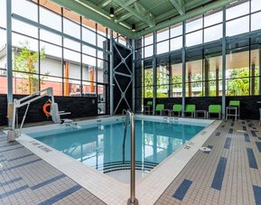 Bright indoor hotel swimming pool surrounded by floor-to-ceiling windows, tiled deck, lounge chairs, accessibility lift, and modern architectural details.