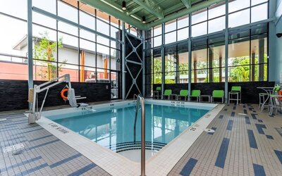 Bright indoor hotel swimming pool surrounded by floor-to-ceiling windows, tiled deck, lounge chairs, accessibility lift, and modern architectural details.