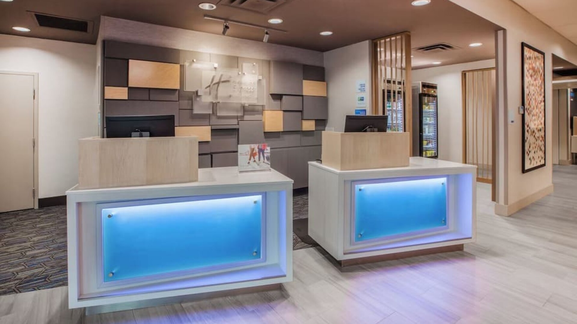 Contemporary hotel reception area with two illuminated front desks, neutral wood finishes, decorative wall panels, and a bright modern lobby.