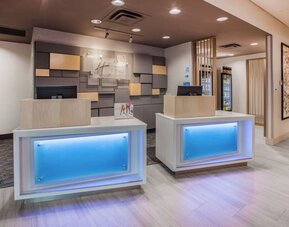 Contemporary hotel reception area with two illuminated front desks, neutral wood finishes, decorative wall panels, and a bright modern lobby.