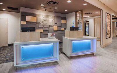 Contemporary hotel reception area with two illuminated front desks, neutral wood finishes, decorative wall panels, and a bright modern lobby.